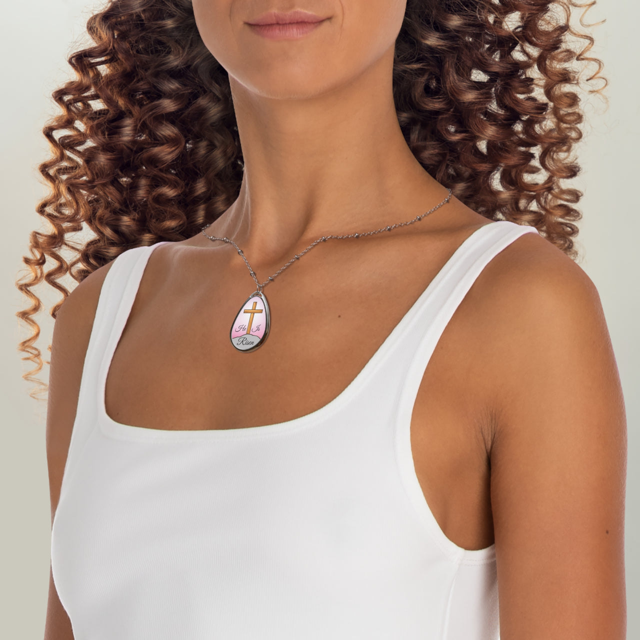 Inspirational oval pendant necklace with "He Is Risen" design,  cross on a hill artwork, zinc alloy and white aluminum, spiritual jewelry for resurrection faith, pink sky, woman wearing necklace
