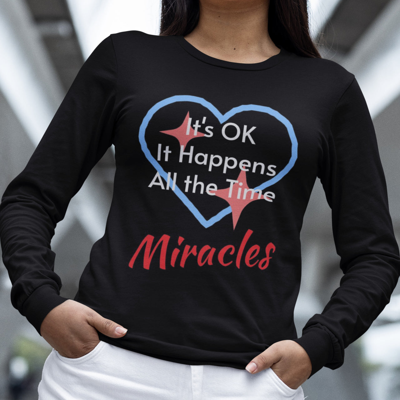 Long-sleeve Bella+Canvas t-shirt made from preshrunk ring-spun cotton with “Miracles Happen” design and message, “It’s OK. It Happens All the Time – Miracles,” faith-based apparel encouraging daily miracles through intimacy with God, woman wearing long-sleeve black t-shirt