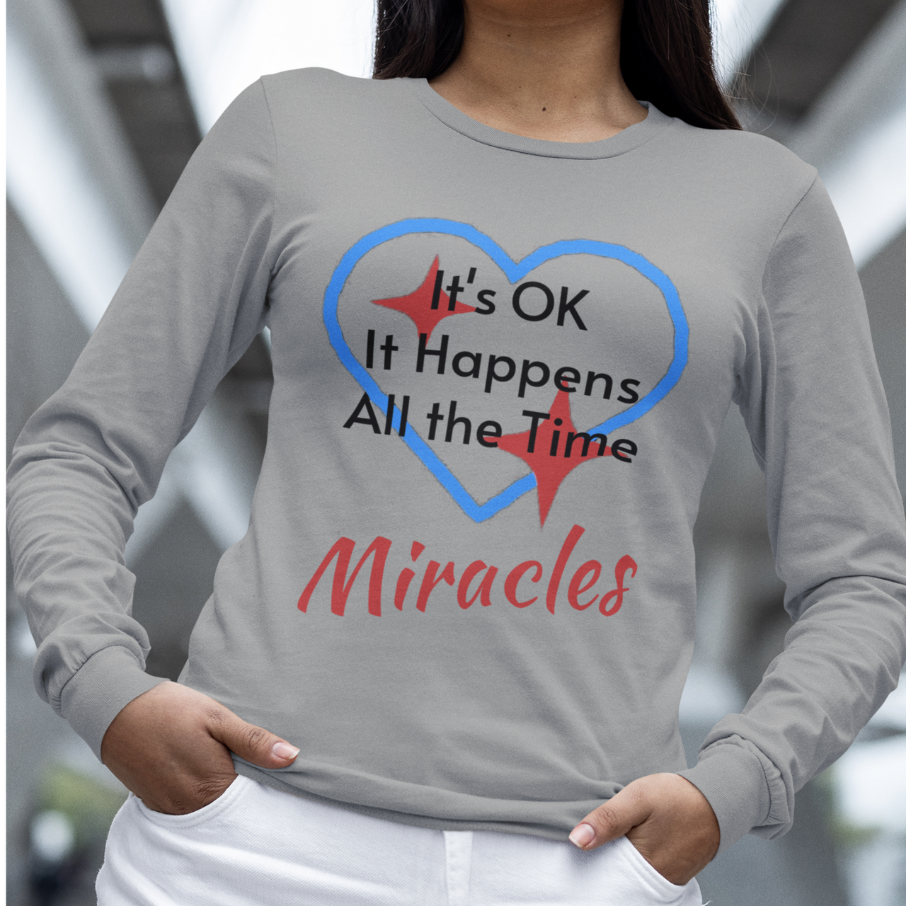 Long-sleeve Bella+Canvas t-shirt made from preshrunk ring-spun cotton with “Miracles Happen” design and message, “It’s OK. It Happens All the Time – Miracles,” faith-based apparel encouraging daily miracles through intimacy with God, woman wearing long-sleeve gray t-shirt