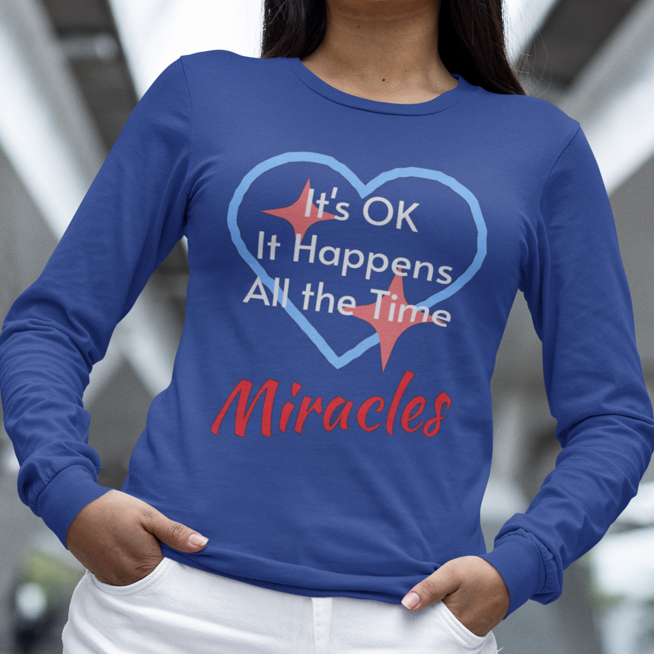 Long-sleeve Bella+Canvas t-shirt made from preshrunk ring-spun cotton with “Miracles Happen” design and message, “It’s OK. It Happens All the Time – Miracles,” faith-based apparel encouraging daily miracles through intimacy with God, woman in long-sleeve blue t-shirt