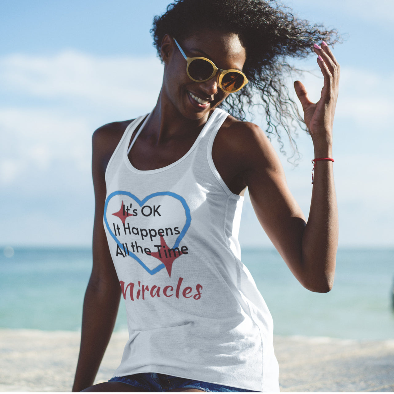 Lightweight Bella+Canvas tank top made from preshrunk cotton with “Miracles Happen” design message, “It’s OK. It Happens All the Time – Miracles,” inspirational apparel encouraging daily miracles through intimacy with God, woman at beach wearing white tank top
