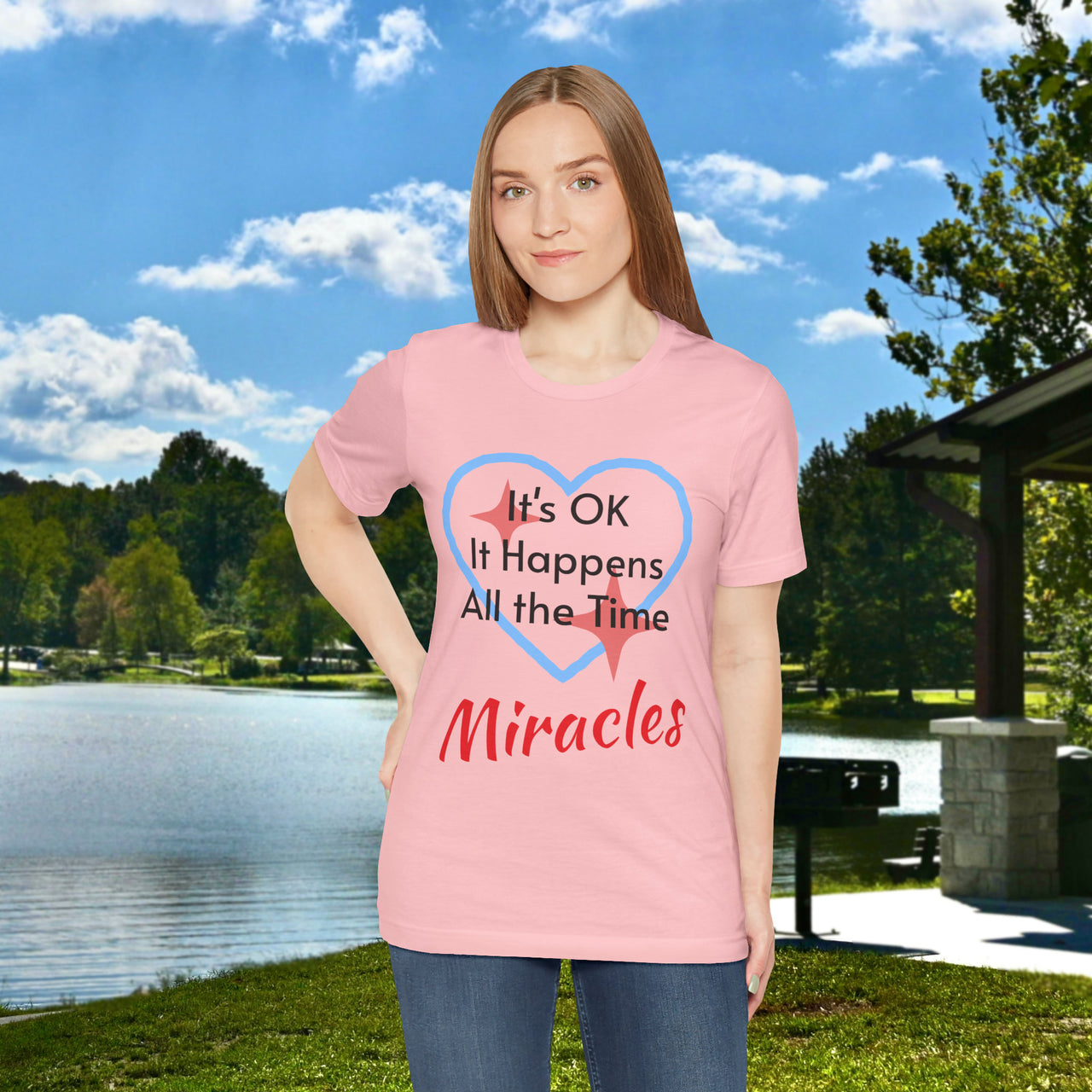 Soft, breathable Bella+Canvas t-shirt made from preshrunk cotton with “Miracles Happen” design message, “It’s OK. It Happens All the Time – Miracles,” inspirational shirt encouraging intimacy with God that produces everyday miracles, woman in pink t-shirt at lakeside park